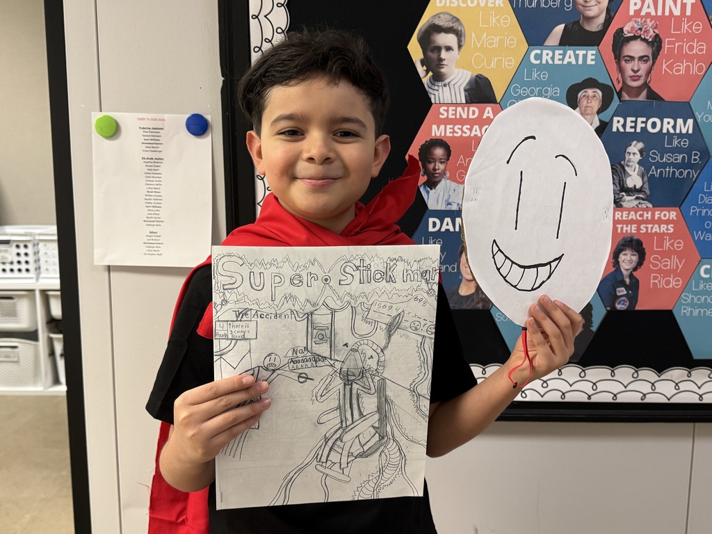 A student dressed as his original comic book character, Super Stickman, smiles while holding his hand-drawn comic book and a mask he created.
