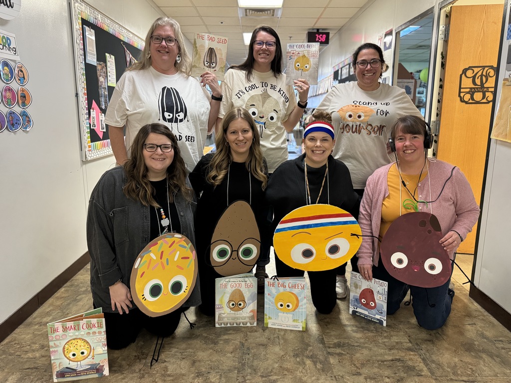 A group of teachers pose in a school hallway wearing themed shirts and holding props and books from “The Food Group” children’s book series including The Bad Seed, The Good Egg, and The Couch Potato.