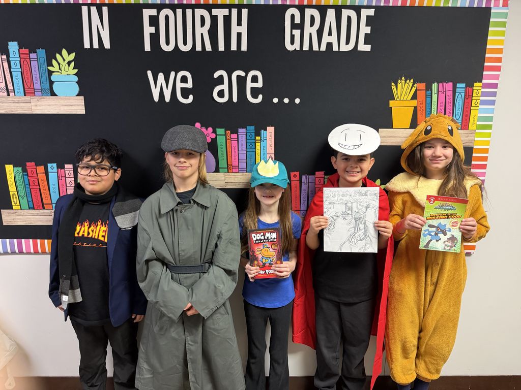 Five students stand in front of a bulletin board that reads “In Fourth Grade We Are…” while dressed as book characters and holding books and drawings, including a student dressed as Super Stickman.