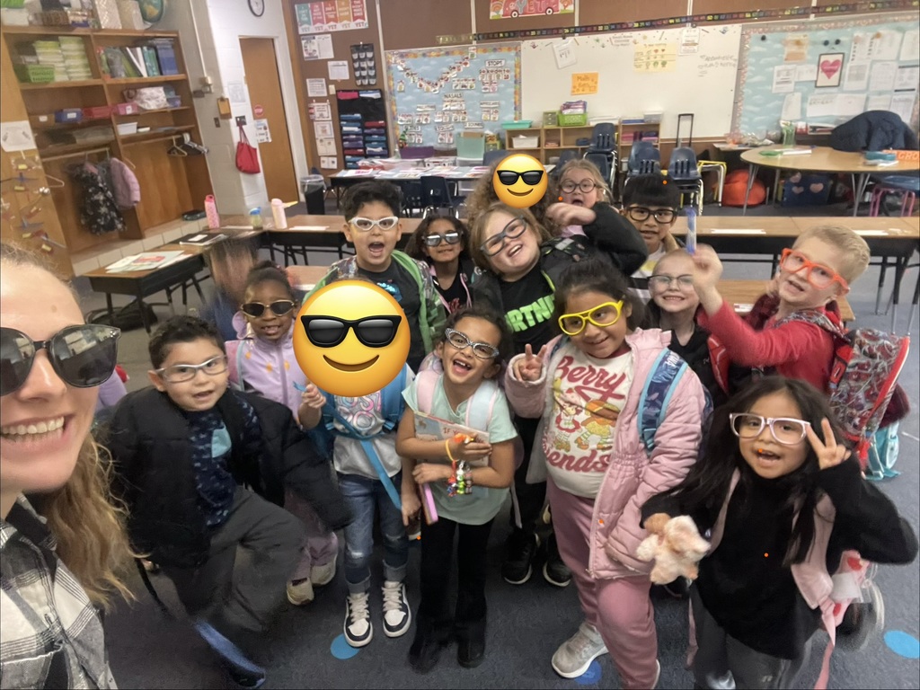 A teacher takes a selfie with a group of young students inside a classroom. The children stand close together smiling, wearing playful glasses and backpacks, while classroom tables, cubbies, and learning materials appear in the background.