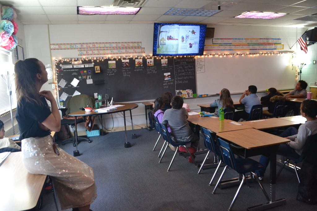 Students sit at desks in a classroom watching a digital screen at the front of the room displaying a picture book. A teacher sits on the front table looking toward the screen, while the classroom walls show alphabet charts, a chalkboard with lessons written on it, and string lights decorating the board.