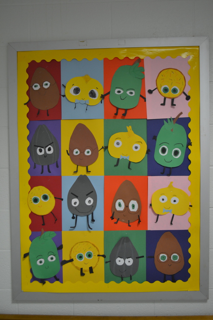A colorful classroom bulletin board displays student artwork inspired by the “Food Group” book series. Paper characters resembling seeds, eggs, grapes, cookies, and beans with cartoon faces are arranged in a grid of bright colored squares and bordered with yellow scalloped trim.