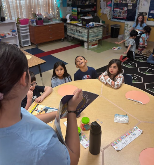 A middle school NJHS student reads aloud from a picture book while sitting at a classroom table with several young elementary students gathered around listening.