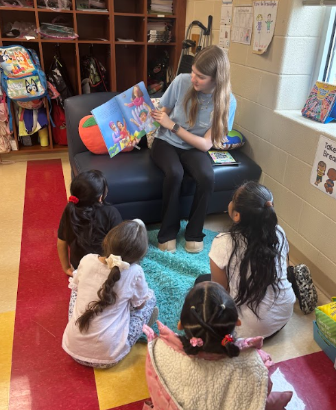 A middle school student from NJHS sits on a small chair in a classroom reading a picture book to a small group of young elementary students seated on the carpet in front of her.