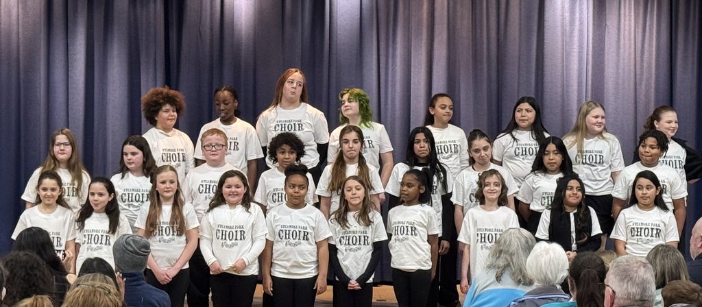A large group of students standing in three rows on a stage with purple curtains. They are all wearing white t-shirts that say "SYCAMORE PARK CHOIR" in black lettering.