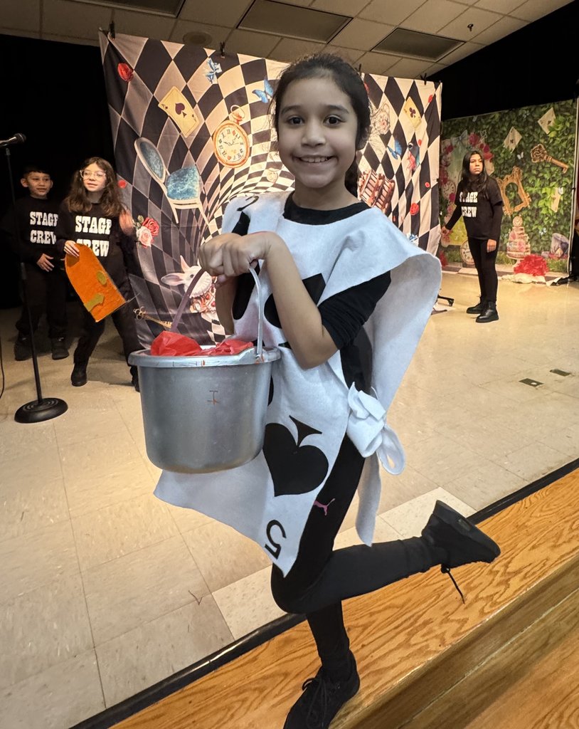A smiling student dressed as a Card Soldier (5 of Spades) poses on one leg on the edge of a stage. In the background, other students in "STAGE CREW" shirts stand near a psychedelic, checkered backdrop.