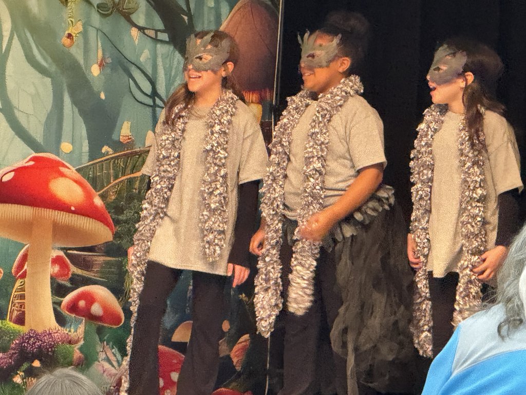 Three children on stage wearing grey tunics, fluffy silver garlands, and dark butterfly-style masks. They are performing in front of a whimsical forest backdrop featuring large red mushrooms.