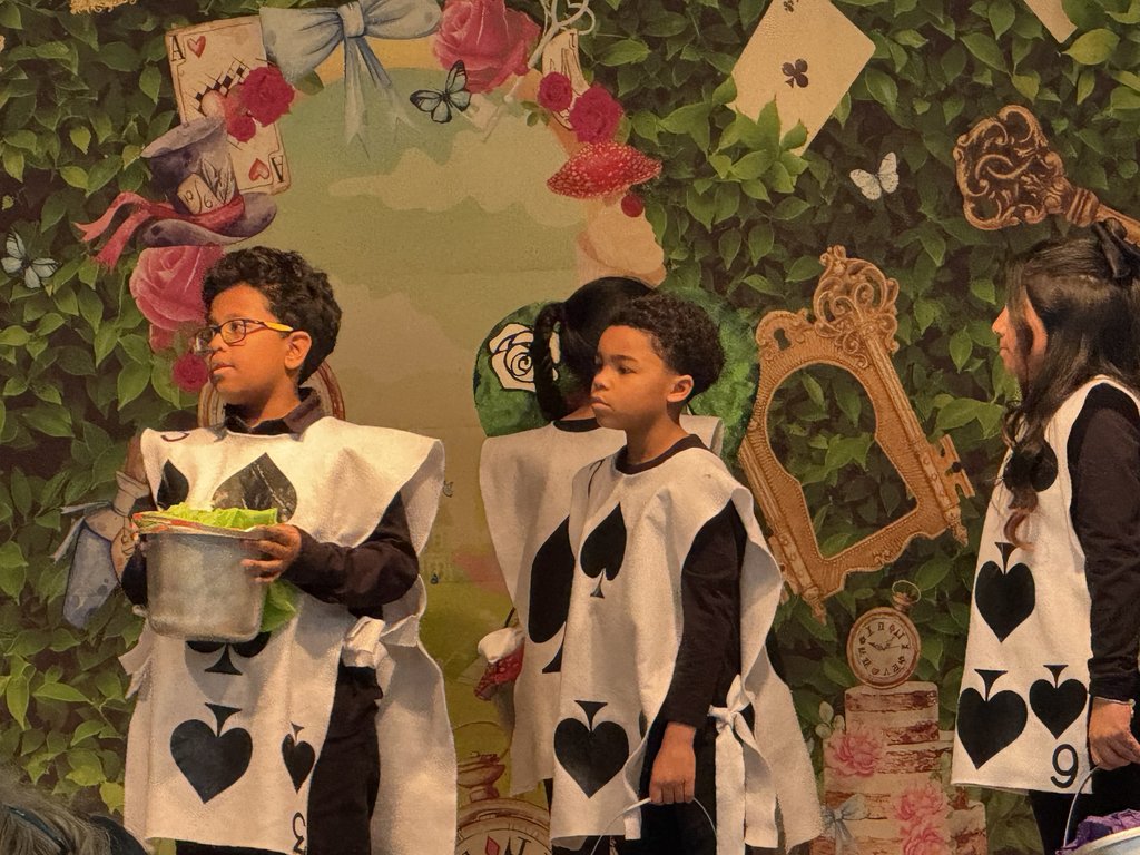 Three students dressed as "Card Soldiers" from Alice in Wonderland. They wear white rectangular tunics with black spade symbols over black long-sleeved shirts and pants.