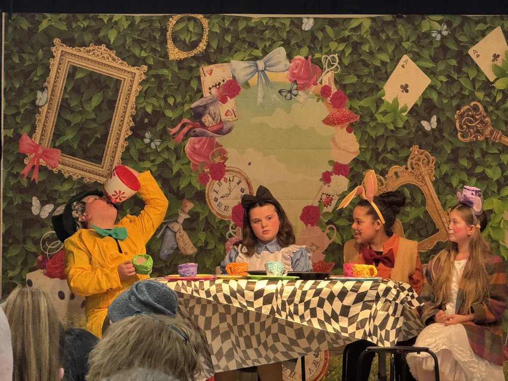 Four children performing the "Mad Tea Party" scene. One child in a yellow coat drinks from a large cup, while others dressed as Alice, the March Hare, and the Mouse sit at a table covered in a checkered cloth.