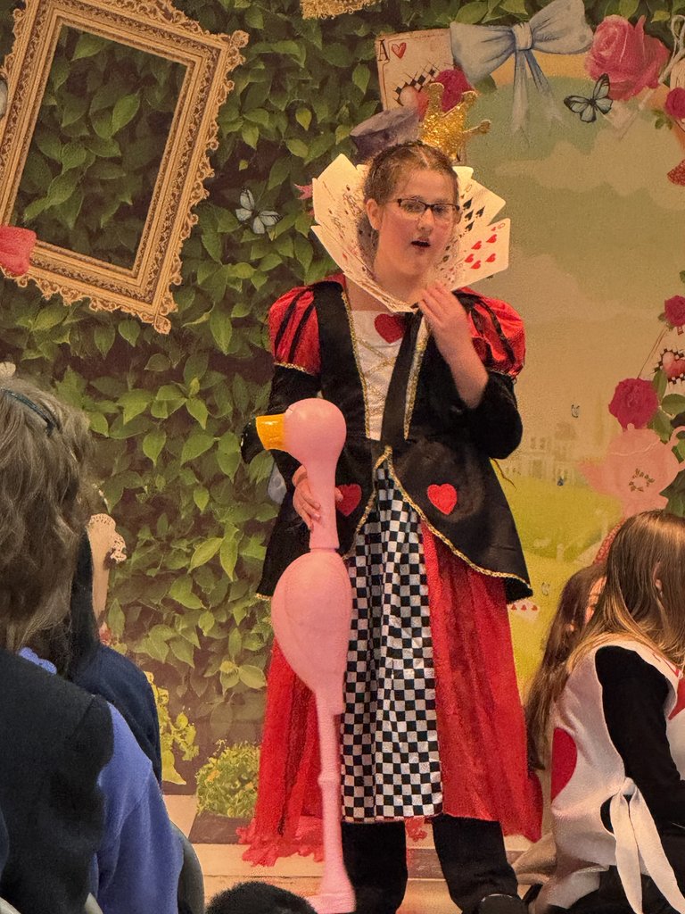 student dressed as the Queen of Hearts wearing a red, black, and gold gown with a high card-themed collar. She is holding a pink plastic flamingo as a mallet.
