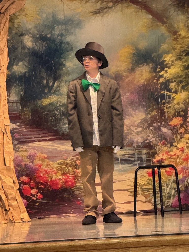 A student dressed as the Mad Hatter stands alone on stage. He is wearing a brown blazer, a large green bowtie, a brown top hat, and round glasses against a painted forest backdrop.