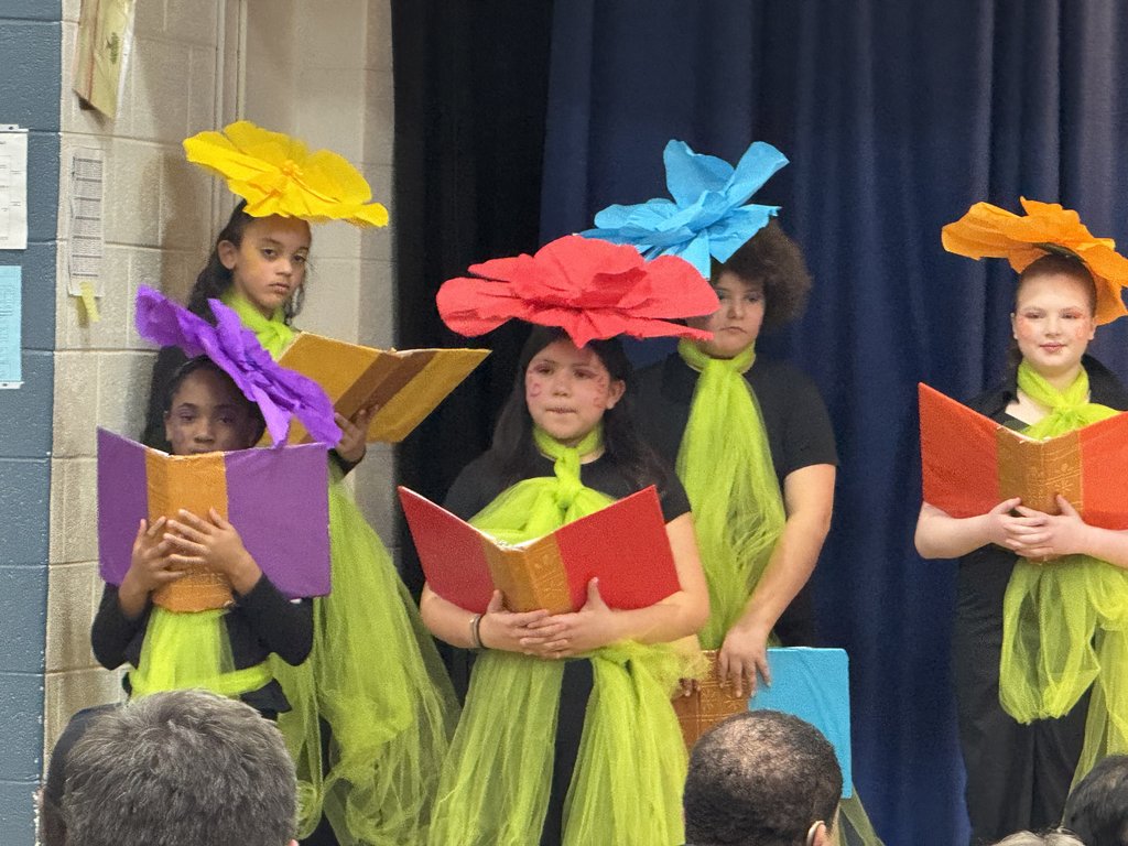 Five children dressed as flowers, wearing bright green mesh wraps and oversized colorful paper flower headpieces in yellow, purple, red, blue, and orange. They are holding open books as if singing.