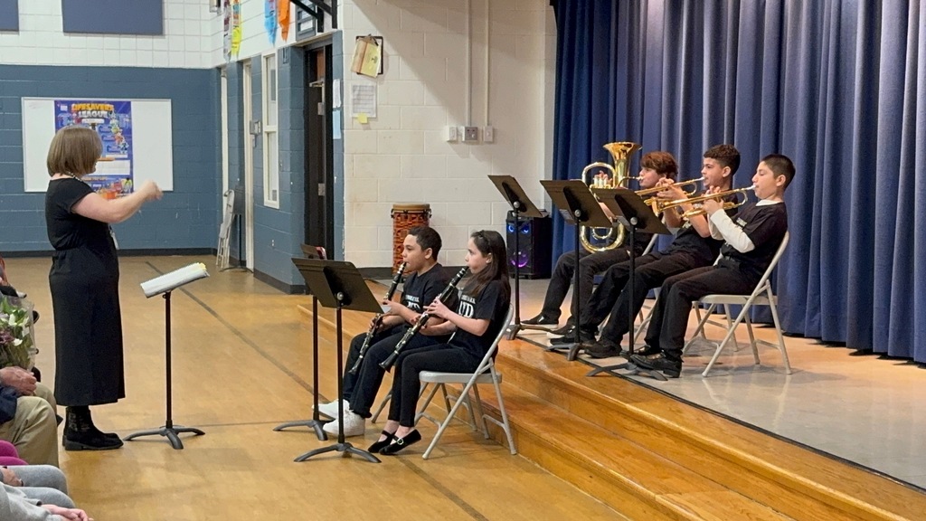 A school band rehearsal in a gymnasium. A conductor leads five students seated on a stage; two play clarinets, two play trumpets, and one plays a tuba.