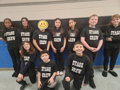 A group photo of eleven students posing against a blue and white wall. They are all wearing matching black t-shirts that say "STAGE CREW" in bold white letters. One student’s face is covered with a yellow smiley face emoji.
