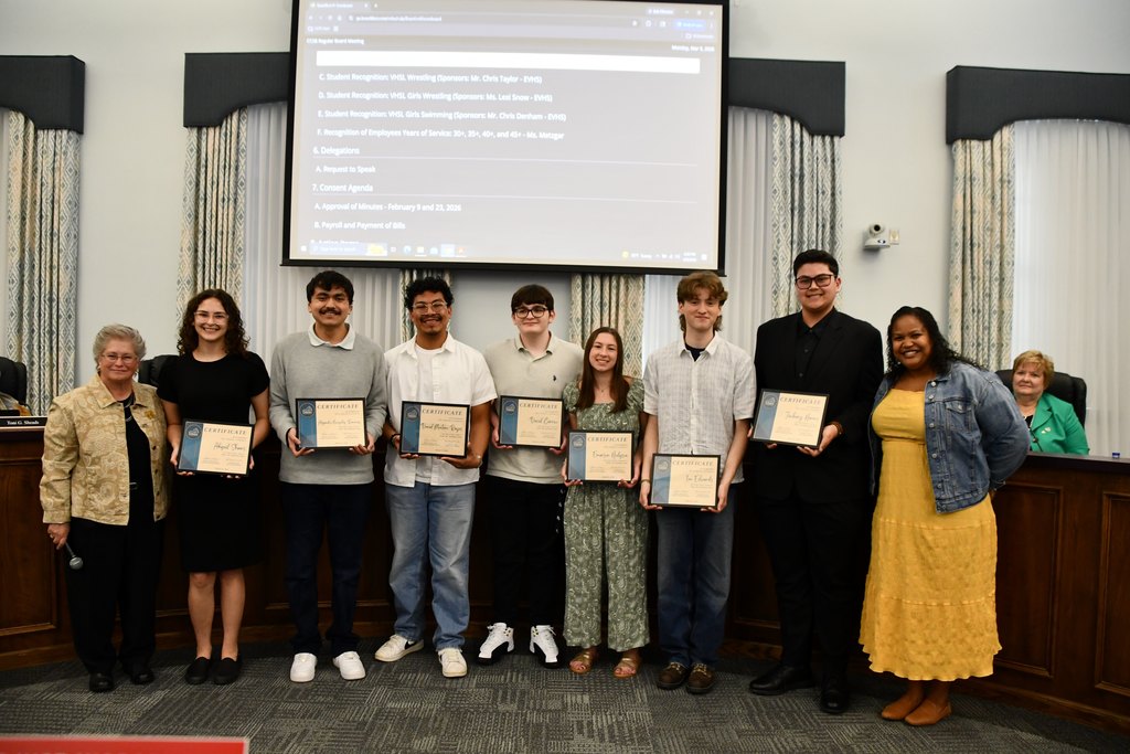 group of seven students holding certificates in a board room with a board member on the left and teacher on the right