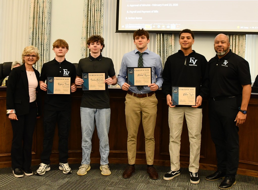 four young men holding certificates in a board room with a board member on the left and their coach on the right