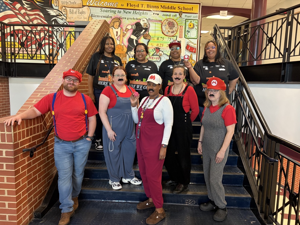 A group of nine adults pose together on a staircase inside a school building decorated with colorful educational murals and a sign reading “Welcome to Floyd T. Binns Middle School.” Several people are dressed as characters inspired by the Mario video game series, wearing red hats with an “M,” overalls, and fake mustaches. Others wear black graphic T-shirts. The group smiles toward the camera, and one person holds a small red mushroom prop. The setting appears to be a school hallway with railings and bright artwork in the background.