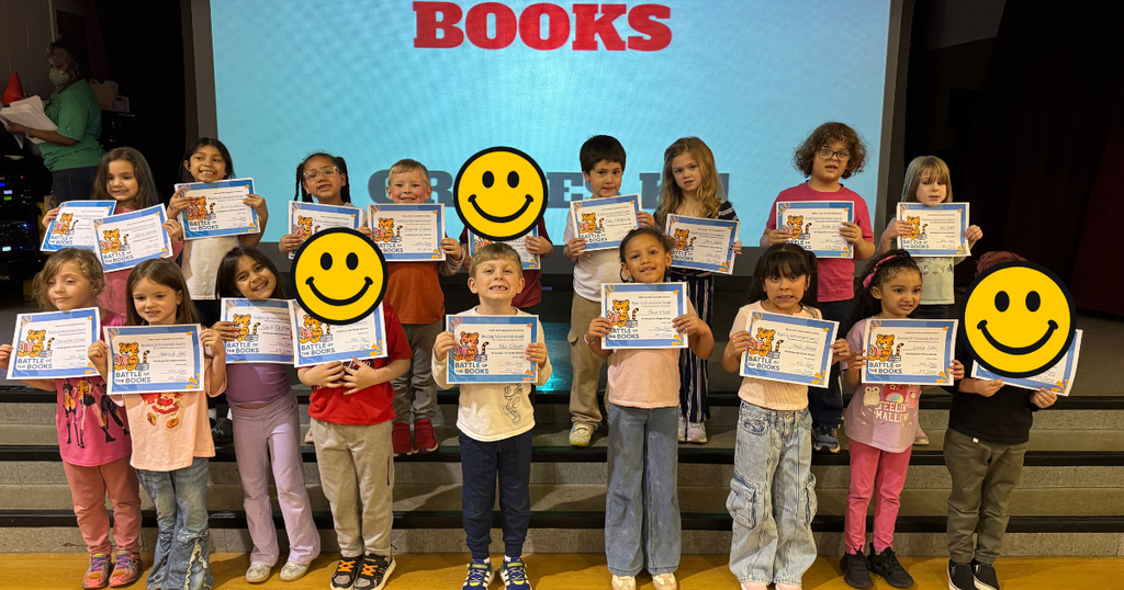 A group of kindergarten students stands on a stage holding “Battle of the Books” certificates in front of a large screen that reads “Battle of the Books.”