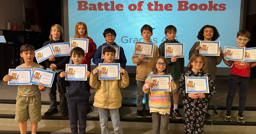 Fourth grade students stand on stage holding their Battle of the Books certificates in front of a presentation screen.
