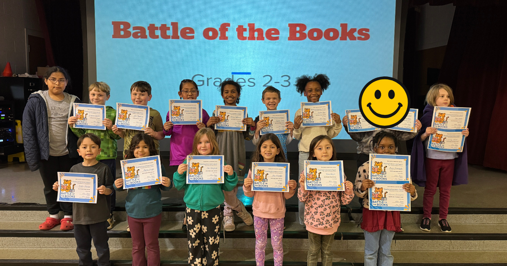 Second grade students stand in two rows on stage proudly holding their Battle of the Books certificates.