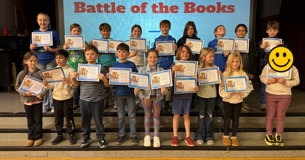 Third grade students pose with their certificates on stage in front of a screen displaying “Battle of the Books – Grades 2–3.”