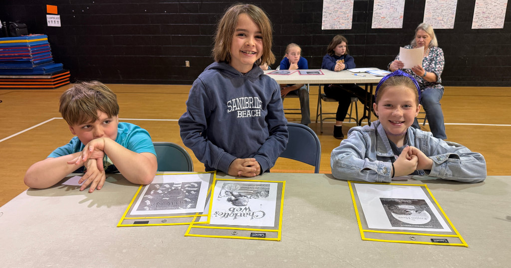 Three students sit at a table with book packets during the Battle of the Books competition while staff members observe in the background.