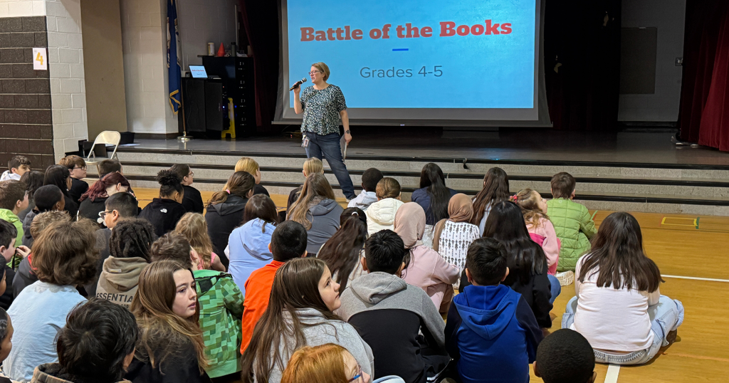 Students sit on the gym floor watching a teacher speak into a microphone in front of a screen that reads “Battle of the Books – Grades 4–5.”