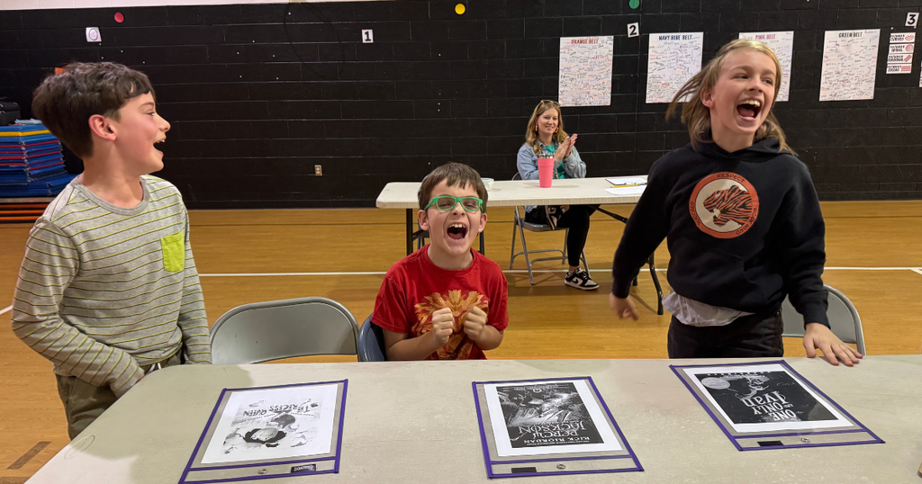 Three students react excitedly during the competition while sitting at a table with book materials.