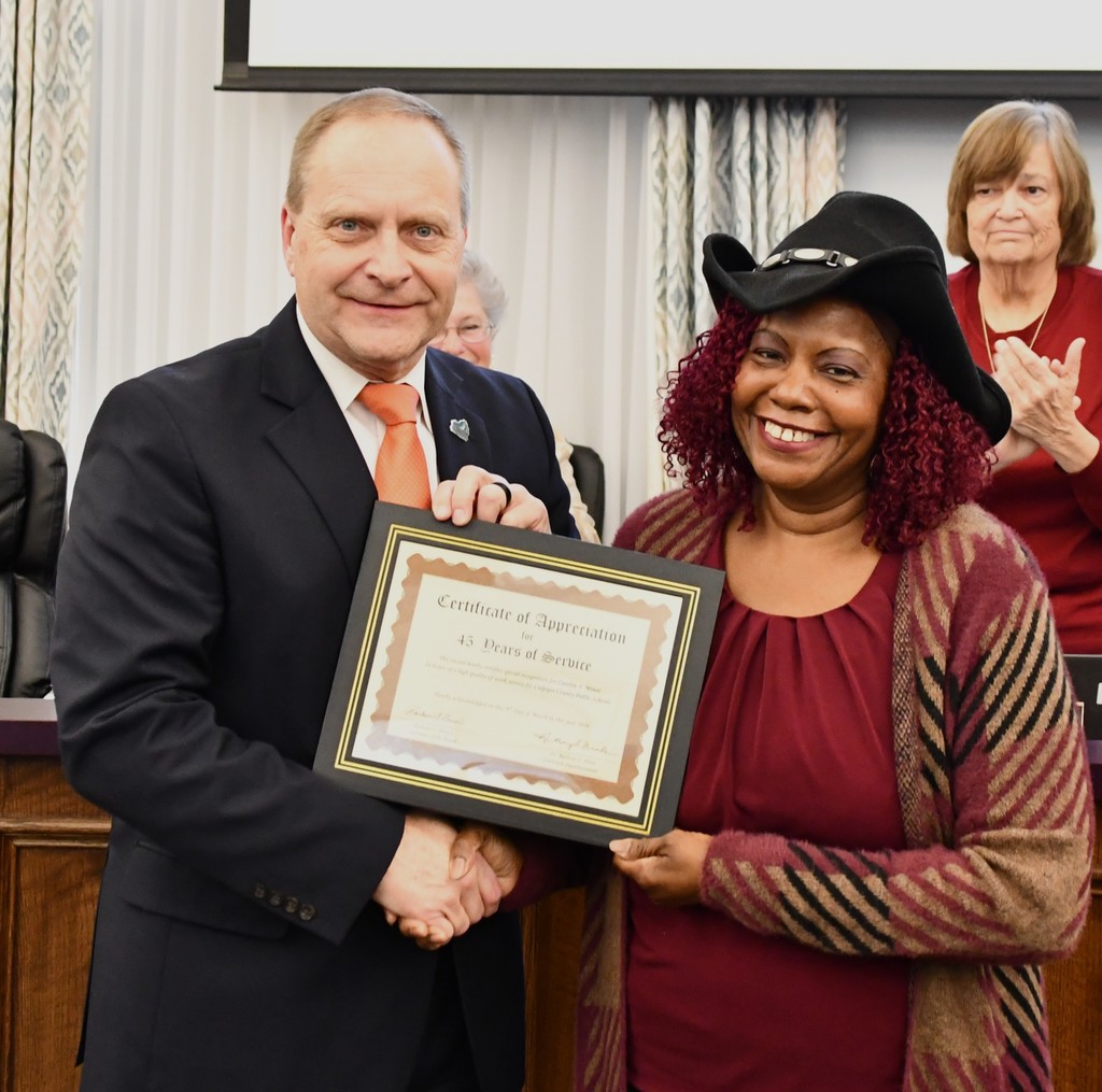 A man in a suit shakes hands with a smiling woman wearing a black cowboy hat as they hold a framed certificate recognizing 45 years of service. They stand in front of a school board dais while several seated board members behind them applaud during the recognition ceremony.