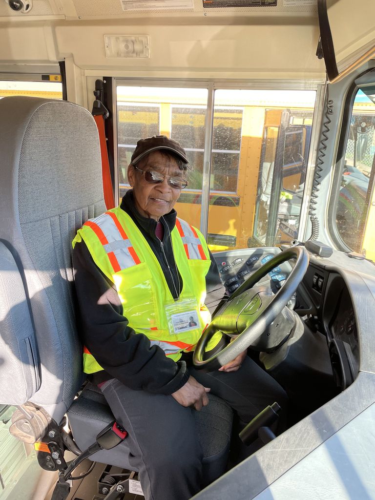 bus driver wearing neon yellow vest sitting in driver seat of a school bus looking at the camera