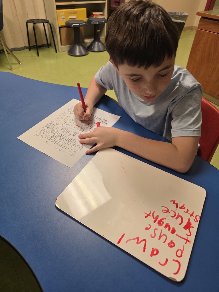 A student sits at a classroom table coloring a birthday cake worksheet with a red crayon while a whiteboard with writing sits beside them.