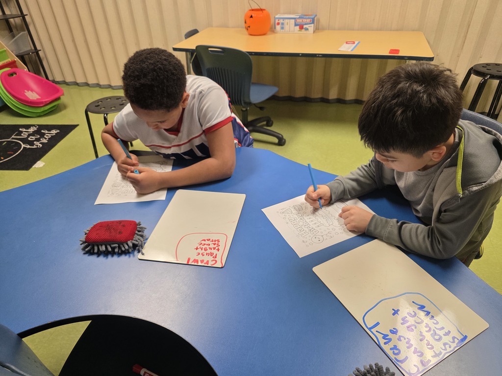 Two children sit at a blue kidney-shaped table coloring birthday-themed worksheets with crayons. Small whiteboards and microfiber erasers are on the table, and classroom furniture and supplies are visible in the background.