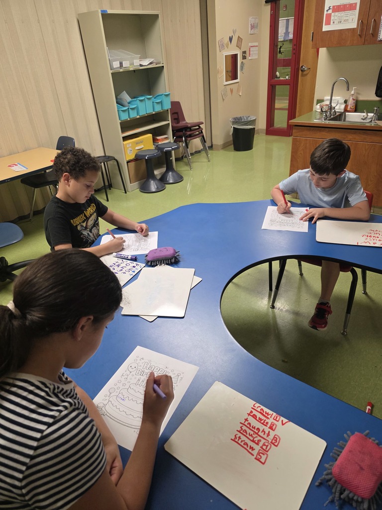 Several students sit around a kidney-shaped classroom table coloring birthday worksheets. Whiteboards with writing and crayons are scattered across the table.