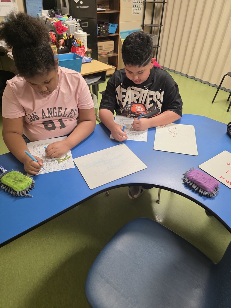 Two students sit together at a blue classroom table focused on coloring birthday worksheets. Whiteboards and cleaning erasers are placed on the table in front of them.