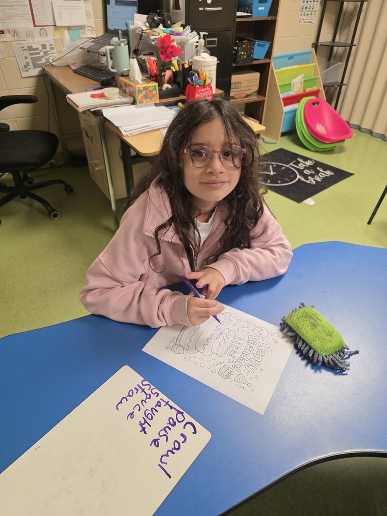 A young student with glasses sits at a classroom table coloring a “Happy Birthday” worksheet with a purple crayon. A whiteboard and cleaning eraser sit nearby, and a teacher’s desk and school supplies are visible behind the student.