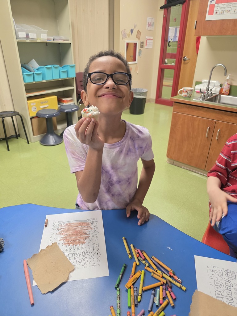 A smiling student holds up a cupcake while standing at a classroom table. Crayons and a partially colored birthday worksheet lie on the table in front of them.