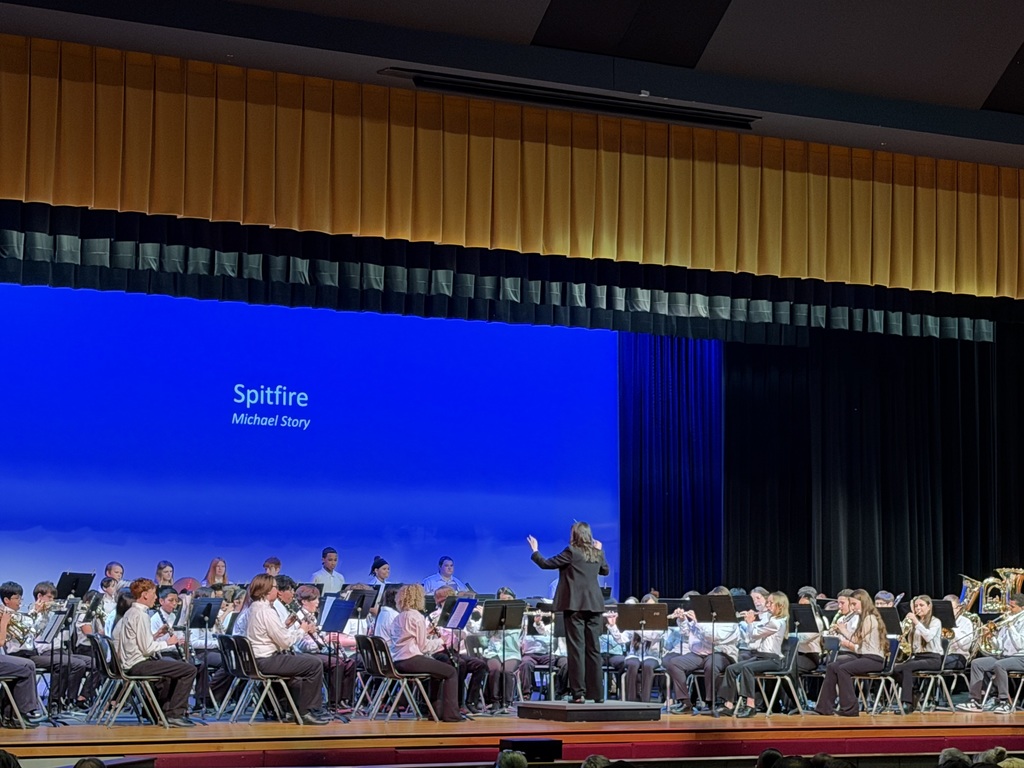 A middle school concert band performs on stage while a conductor stands on a podium directing the musicians. Students seated in rows play a variety of instruments including flutes, clarinets, trumpets, and tubas. A large screen behind them displays the title “Spitfire” by Michael Story against a blue background, and audience members are visible watching from the foreground.
