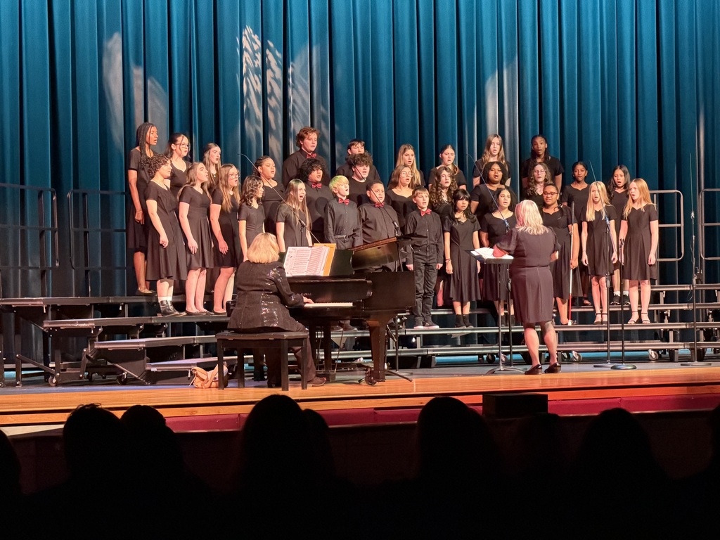 A middle school choir performs on stage in front of tall teal curtains, standing on tiered risers in black concert attire. Some students wear red bow ties while others wear black dresses. A conductor stands in front of the group directing the performance, while a pianist plays a grand piano beside the risers. Audience silhouettes are visible in the foreground.