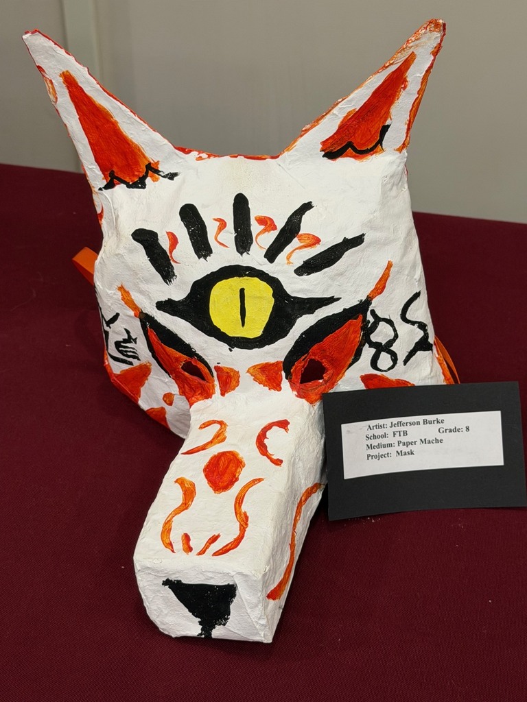 A stylized paper mache animal head mask painted white with orange and black markings and a large yellow eye on the forehead, displayed with a student label.