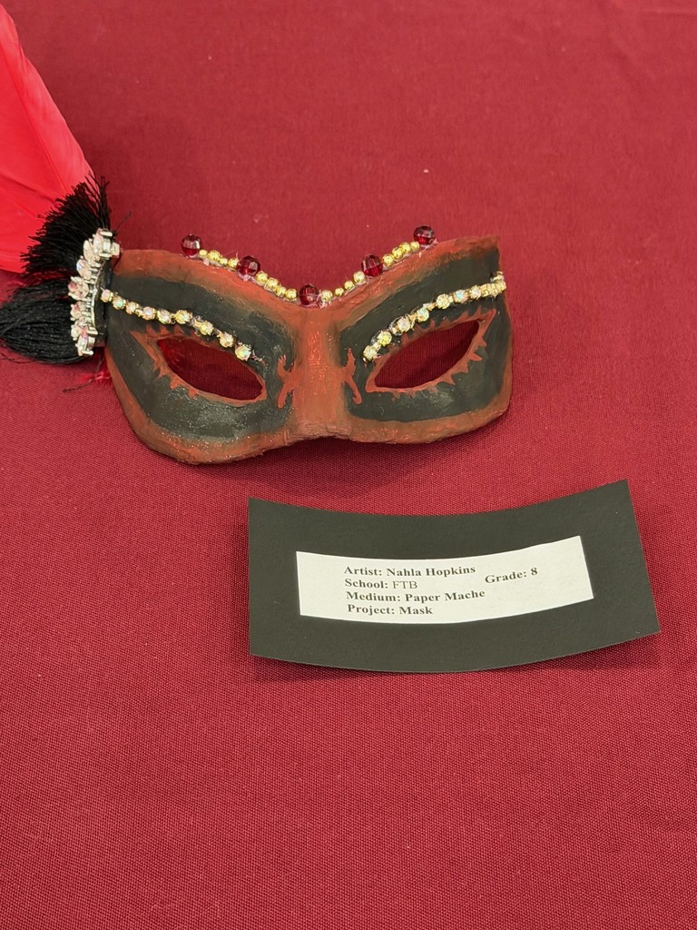 A decorative mask painted in black and red with gold accents and small jewels, featuring a tassel detail on one side. A label card below lists the student artist and project.