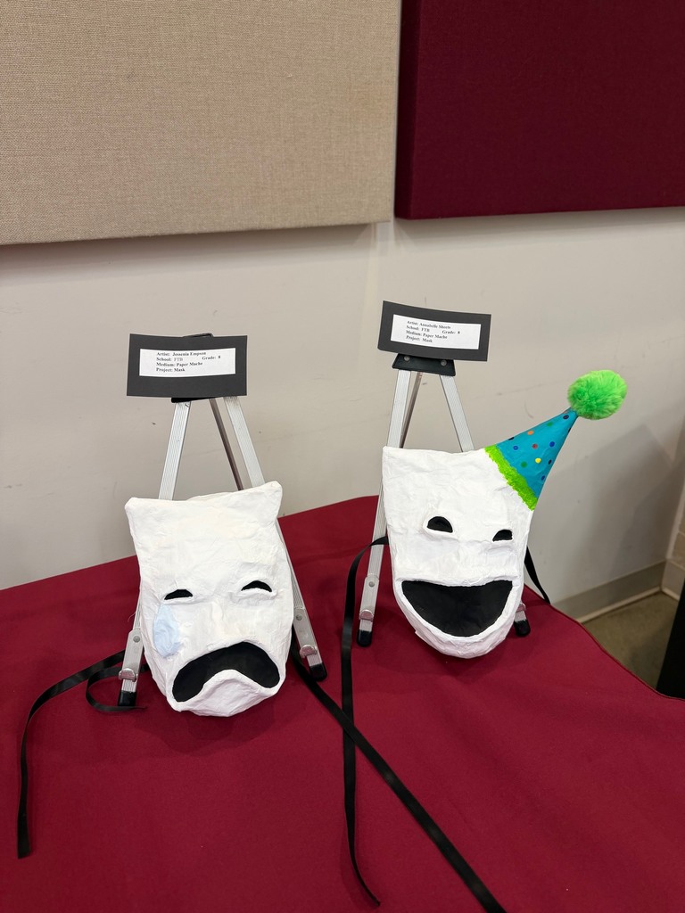 Two white paper mache masks displayed on small easels, one with a sad expression and the other with a smiling face wearing a blue party hat with a green pom-pom. Both are labeled as student artwork.