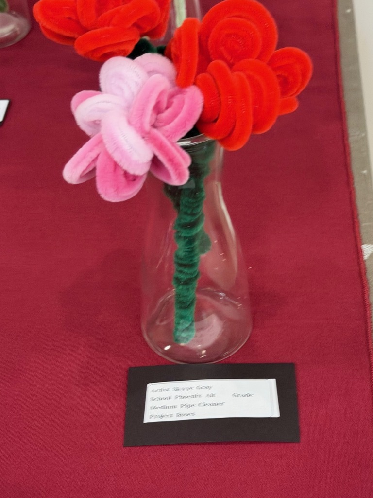 A small bouquet of handmade flowers crafted from colorful pipe cleaners—red and pink blossoms with green stems—displayed in a clear glass bottle on a red tablecloth. A label card sits in front identifying the student artwork.