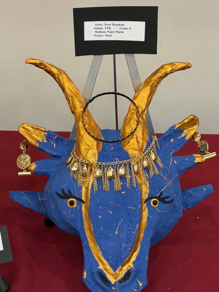 A sculptural paper mache piece shaped like a blue animal head with large gold horns, gold accents, and decorative jewelry elements, displayed on a stand with a label above it.
