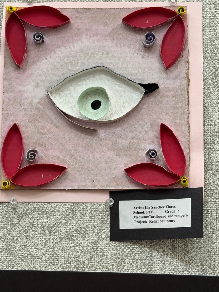 A mixed-media relief sculpture featuring a central eye surrounded by red leaf-like shapes and curled elements, mounted on a board with a label identifying the student and medium.