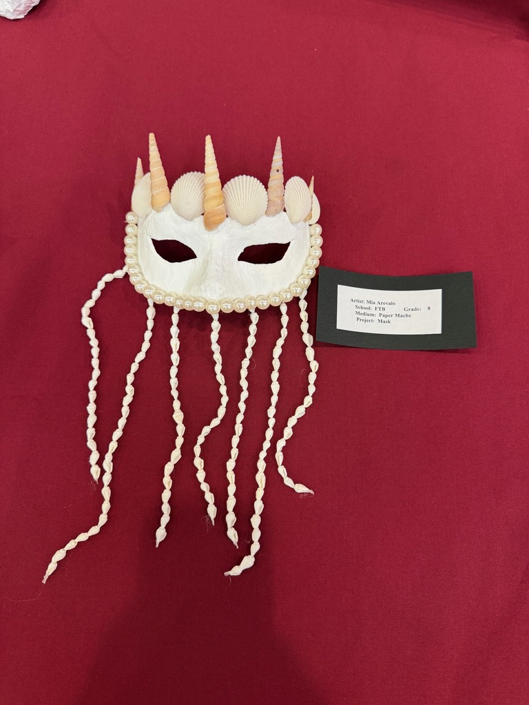 A white paper mache mask decorated with seashells and strands of beads hanging down, giving it an ocean-inspired look. A label card identifies the student artist and project.