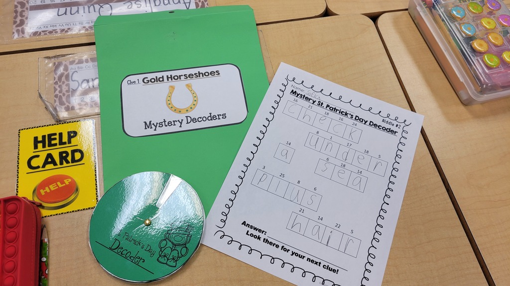 A St. Patrick’s Day activity setup on a desk includes a “Help Card,” a spinner labeled “Decoders,” and a worksheet where a student has decoded a message reading “Check under a plush chair.”