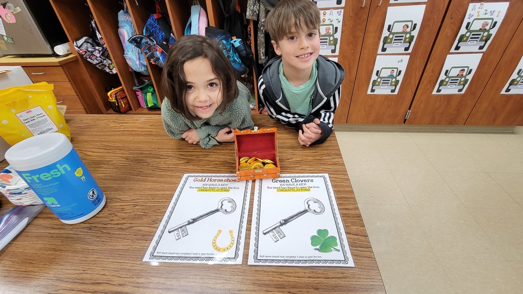 Two students lean over a classroom table smiling next to a small treasure chest filled with gold coins. Printed clue sheets with keys, a horseshoe, and a clover are laid out in front of them.