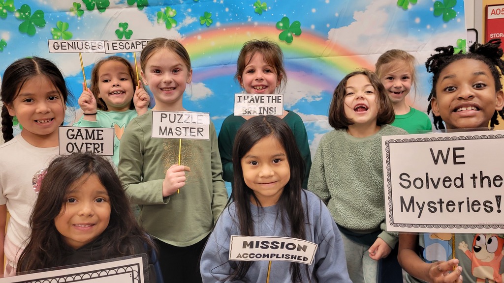 A group of smiling students pose in front of a rainbow and shamrock backdrop holding signs that say phrases like “Puzzle Master,” “Game Over,” and “We Solved the Mysteries!”