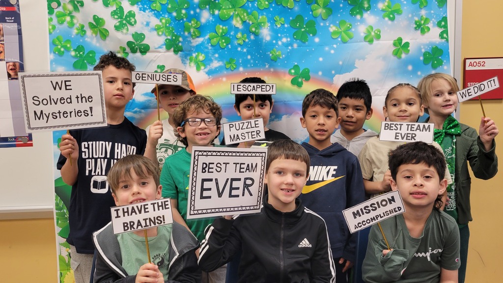 A group of elementary students stand in front of a rainbow and shamrock-themed backdrop, smiling and holding signs that read messages like “We Solved the Mysteries,” “Best Team Ever,” “Mission Accomplished,” and “I Have the Brains.”