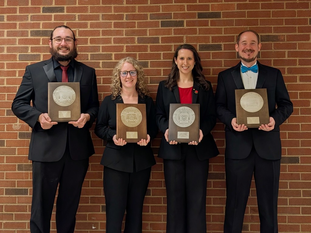 Four CCPS band directors stand in front of a brick wall, each holding a wooden plaque with a Virginia seal, representing Superior ratings earned at the District Band Assessment. They are dressed in formal black concert attire, smiling and posed side by side.